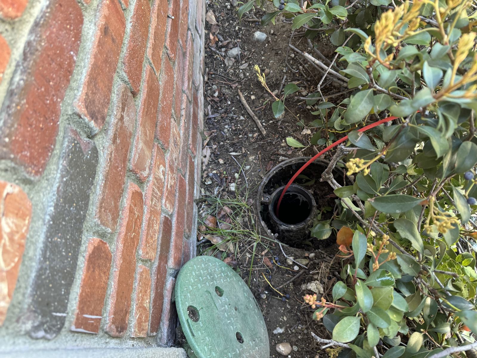 Camera cable entering a ground-level sewer cleanout beside a brick wall