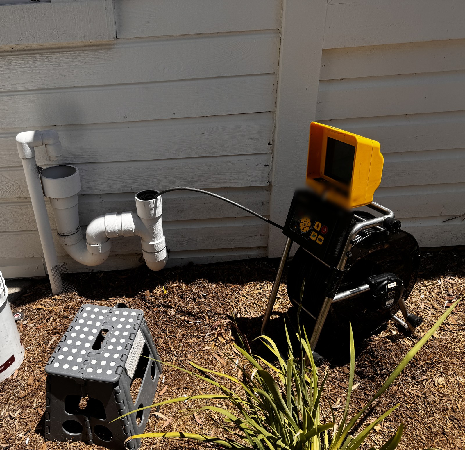 Sewer camera and monitor set up at a side-yard cleanout with exposed PVC piping alongside a residential wall